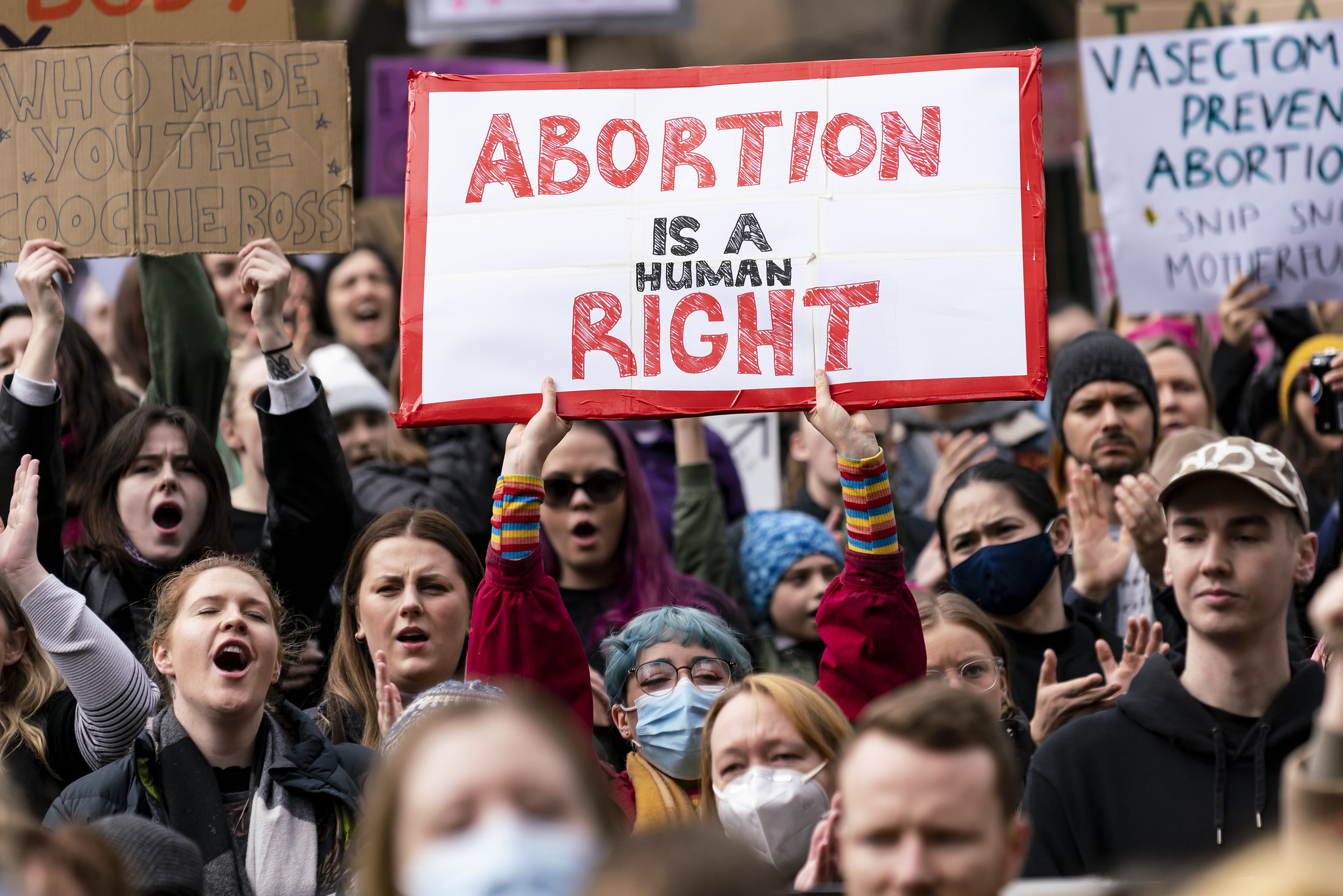 An image from an abortion rights protest. The camera is focused on one person who is wearing a rainbow striped long sleeve top and holding a sign which reads in red 'abortion is a human right'