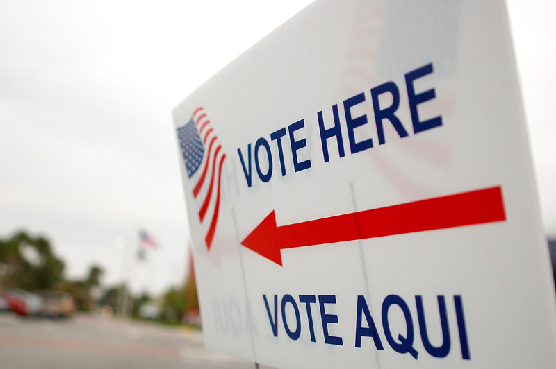 A sign with a red arrow that says "Vote here" and "vote aqui" underneath. There is an American flag in the upper left corner of the sign.